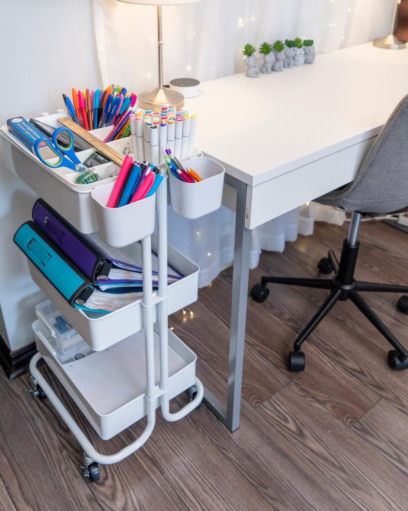 A white three tier storage cart filled with office supplies and binders sits next to a clean white desk. 