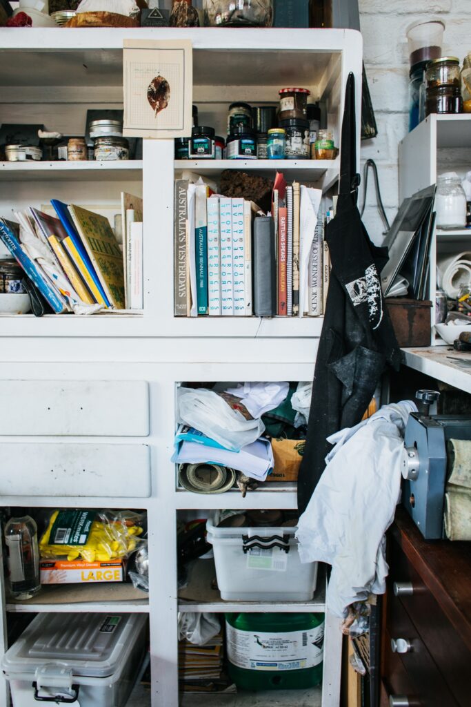 A cabinet in a workshop overflows with books, paints, storage totes, and other supplies. 