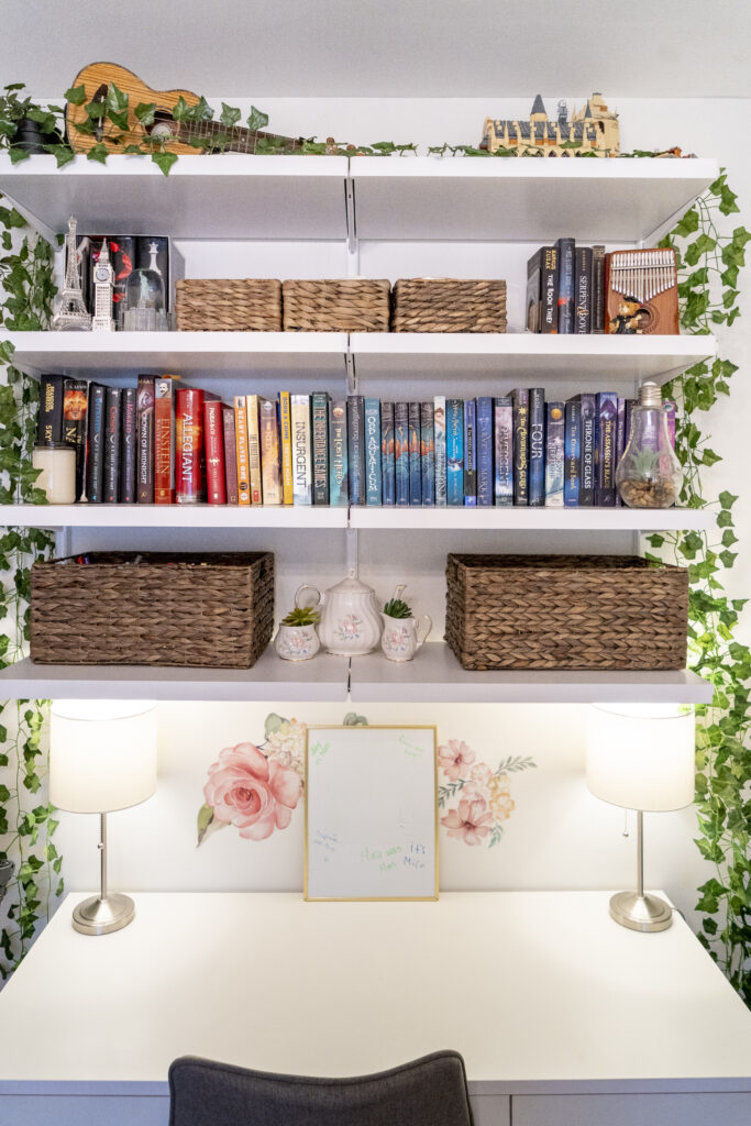 Floating white wall shelves above a desk hold books, wicker baskets, a decorative porcelain tea set, ivy, and other decor pieces.
